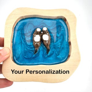 Op de afbeelding: Een houten dienblad met een blauw hars rivierontwerp, met twee bruine otters met witte vlekken. De lade heeft een lichte houten rand en de tekst "Your Personalization" onderaan.
