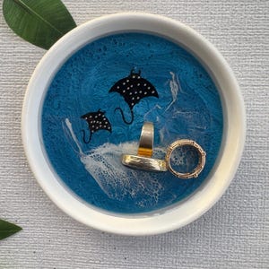May include: A round, white-rimmed trinket dish with a blue resin interior featuring two black manta ray silhouettes. Two rings rest inside the dish. The dish is on a light gray surface.