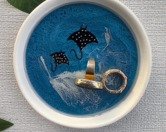 May include: A round, white-rimmed trinket dish with a blue resin interior featuring two black manta ray silhouettes. Two rings rest inside the dish. The dish is on a light gray surface.
