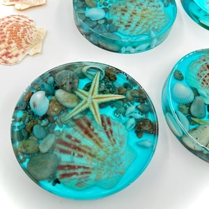 May include: Circular resin coasters with a turquoise blue base, incorporating seashells, pebbles, and starfish. The coasters have a clear, glossy finish. Several are visible, alongside loose seashells.