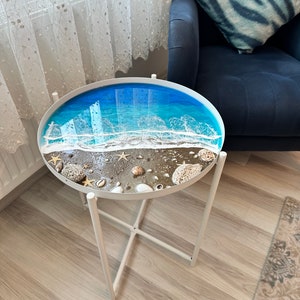 Könnte beinhalten: Ein weißer Beistelltisch aus Metall mit einer ovalen Tischplatte, die eine Strandszene zeigt. Die Tischplatte hat ein blaues Ozean-Design mit weißen Wellen, Sand, Muscheln und Seesternen. Der Tisch hat ein schlichtes, modernes Design.