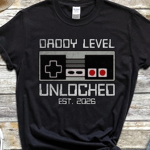 May include: Black t-shirt with the text "DADDY LEVEL UNLOCHED EST. 2026" and a controller graphic. The shirt is surrounded by a pair of black and white sneakers, denim shorts, and a straw hat.