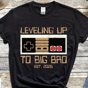 May include: Black t-shirt with a retro video game controller graphic and the text "LEVELING UP TO BIG BRO EST. 2026". The shirt is paired with blue denim shorts and black sneakers.