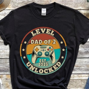 Op de afbeelding: Zwart T-shirt met een vintage-stijl grafische afbeelding met de tekst "Level Dad of 2 Est. 2025 Unlocked" en een retro videogamecontroller.