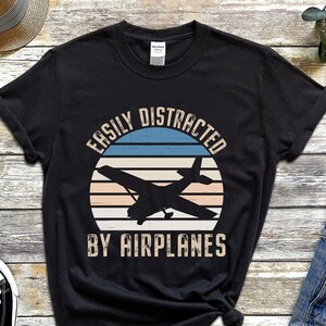 May include: Black t-shirt featuring a vintage-style graphic of an aeroplane in front of a sunset. The text "Easily Distracted By Airplanes" is printed on the shirt.