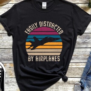 May include: Black t-shirt featuring a vintage-style sunset graphic and the text "Easily Distracted by Airplanes".