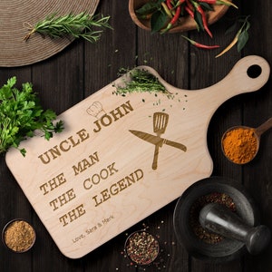 May include: A wooden cutting board with a handle, engraved with the text "Uncle John The Man The Cook The Legend" and a chef's hat and crossed kitchen utensils. The text "Love, Sara & Mark" is also engraved on the board.