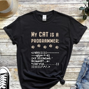 Pode incluir: Camiseta preta com o texto "MY CAT IS A PROGRAMMER:" e uma linha de código abaixo. O texto é de cor marrom claro. Há cinco pegadas de patas acima do texto.