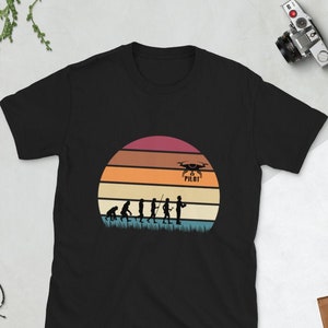 May include: Black t-shirt with a vintage-style sunset graphic featuring a silhouette of the evolution of man culminating in a person holding a drone controller. The text "Drone Pilot" is above the drone.
