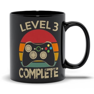 May include: Black ceramic coffee mug with a retro-style design featuring a video game controller and the text "Level 3 Complete".