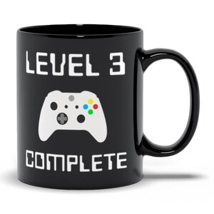 May include: Black coffee mug with a white gamer controller graphic and the text "LEVEL 3 COMPLETE".