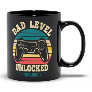 Puede incluir: Taza de café de cerámica negra con un diseño retro que presenta un mando de videojuego y el texto "Dad Level Unlocked Est. 2024".