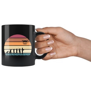 May include: Black coffee mug featuring a white silhouette of the evolution of man, culminating in a person holding a drone with the text "Drone Pilot". The background is a colourful sunset.