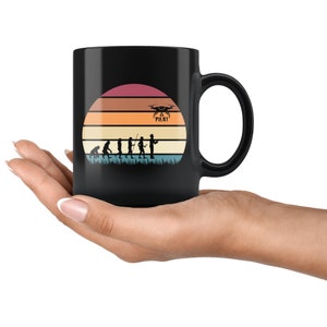 May include: Black coffee mug with a retro sunset design featuring a silhouette of a person holding a drone with the text "Drone Pilot". The design also includes a series of silhouettes depicting the evolution of humans.