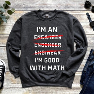 Può includere: Una felpa grigia scura con il testo "I'm an Enganeer Engineer Enginear I'm good with math" stampato sul davanti. Le parole "Enganeer", "Engineer" e "Enginear" sono barrate con linee rosse.
