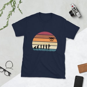 May include: Navy blue t-shirt with a vintage-style illustration of the evolution of man culminating in a drone pilot holding a remote control. The illustration is set against a sunset background with the text "Drone Pilot" above the pilot.