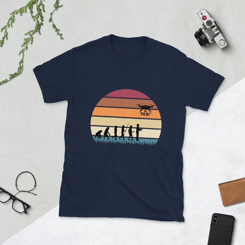 May include: Navy blue t-shirt with a vintage-style illustration of the evolution of man culminating in a drone pilot holding a remote control. The illustration is set against a sunset background with the text "Drone Pilot" above the pilot.