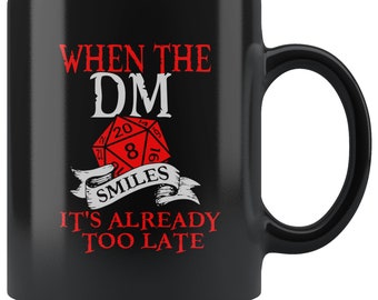 May include: Black ceramic coffee mug with a white design that says "When the DM smiles it's already too late". The design also includes a red 20-sided die.