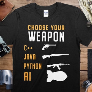 Puede incluir: Camiseta negra con texto blanco que dice "Elige tu arma" y enumera los lenguajes de programación C++, Java, Python e IA con siluetas de armas correspondientes.