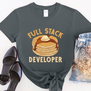 K&ouml;nnte beinhalten: Ein dunkelgraues T-Shirt mit dem Text "Full Stack Developer" und einer Grafik eines Stapels Pfannkuchen mit Butter und Sirup.