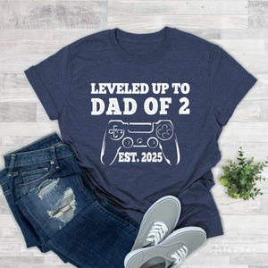 May include: Navy blue t-shirt with the text "LEVELED UP TO DAD OF 2" and "EST. 2025" with a white controller graphic. The shirt is paired with blue jeans and gray sneakers.