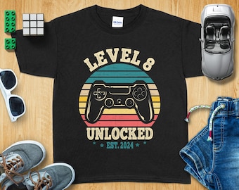 Peut inclure: T-shirt noir avec un graphisme de manette de jeu vidéo rétro et le texte "Level 8 Unlocked Est. 2024".
