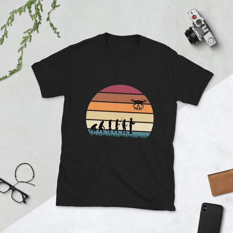 May include: Black t-shirt with a vintage-style sunset graphic featuring a silhouette of the evolution of man culminating in a person holding a drone controller. The text "Drone Pilot" is above the drone.