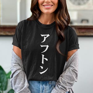 May include: Black t-shirt with white Japanese characters. The shirt has short sleeves and a crew neck. The text is vertically aligned. The person is wearing a gray cardigan and blue jeans.