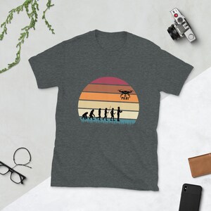 May include: A dark heather grey t-shirt with a vintage-style illustration of the evolution of man, culminating in a person holding a drone controller. The illustration is set against a sunset background with the text "Drone Pilot" above the drone.