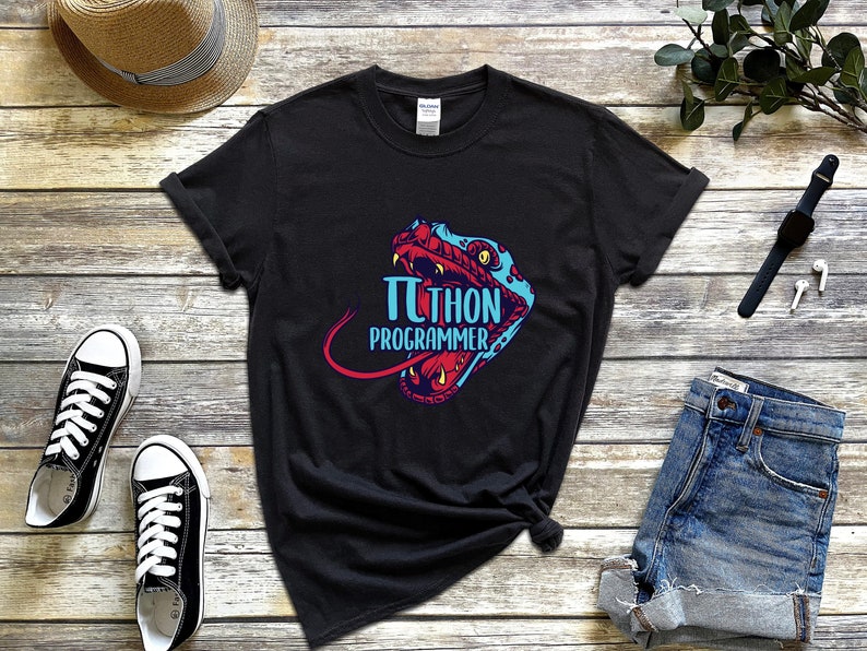Op de afbeelding: Zwart t-shirt met een kleurrijk ontwerp van een slangenkop en de woorden "&pi;THON PROGRAMMER" in blauw en rood. Het shirt wordt getoond met een spijkerbroek, sneakers, een horloge en een hoed op een houten oppervlak.