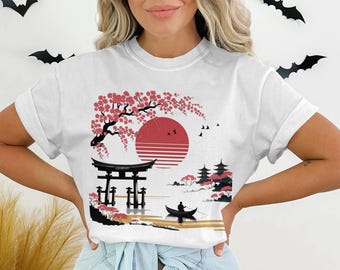 Pode incluir: Camiseta branca com um design de paisagem japonesa. O gráfico inclui um sol vermelho, flores de cerejeira, um portão tradicional, pagodes e uma pessoa em um barco. O design é em vermelho, preto e branco.
