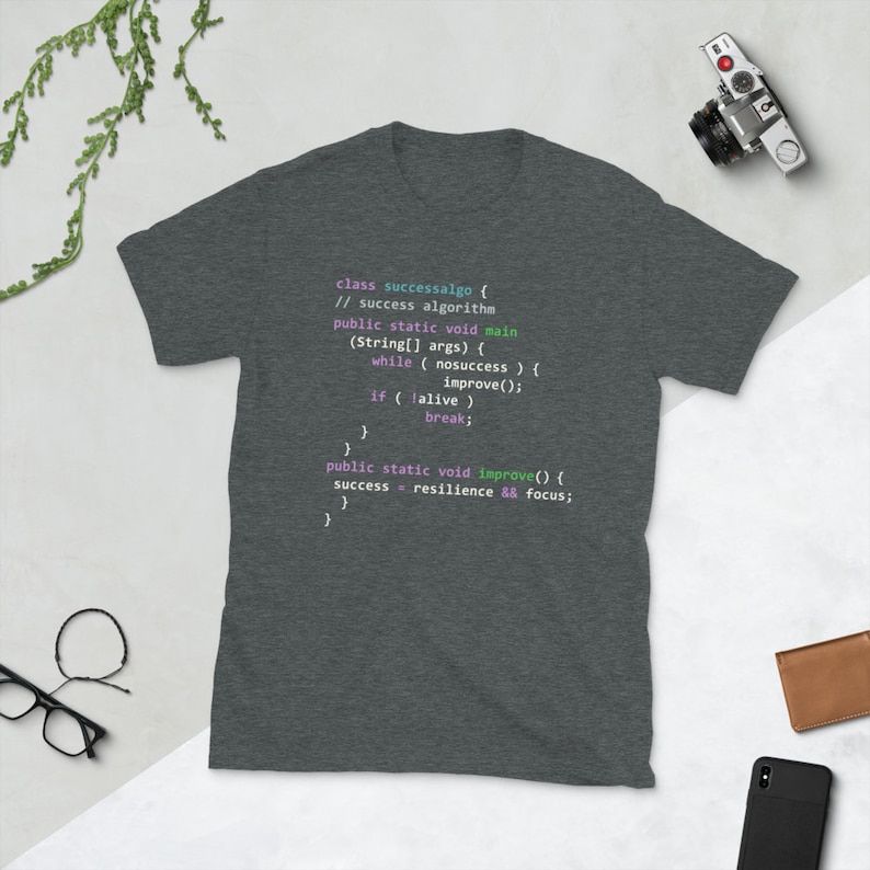 Op de afbeelding: Een donker grijs t-shirt met een code-snippet erop gedrukt. De code is geschreven in lichtblauw, roze en groen. De code luidt: "class successalgo { // success algorithm public static void main (String[] args) { while (nosuccess) { improve(); if (lalive) break; } } public static void improve() { success = resilience && focus; } "