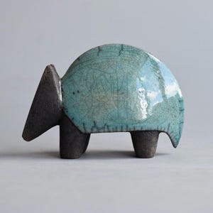 May include: A ceramic sculpture of an animal with a rounded, turquoise-colored back and a dark gray head and legs. The turquoise surface has a crackle glaze. The sculpture is on a light gray background.