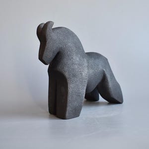 May include: A dark gray, textured sculpture of a horse standing in a profile position. The figure features a curved neck, a solid body, and four legs, with a rough, granular surface finish throughout the entire form of the animal.
