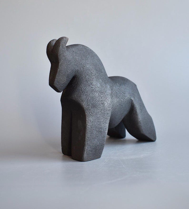May include: A dark gray, textured sculpture of a horse standing in a profile position. The figure features a curved neck, a solid body, and four legs, with a rough, granular surface finish throughout the entire form of the animal.