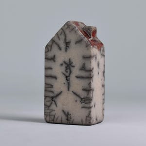 May include: A small, white ceramic house with a red roof and black markings. The house has a textured surface and a slightly uneven shape.