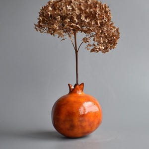 May include: A ceramic pomegranate vase with a glossy orange finish, holding a dried brown hydrangea flower. The vase has a textured surface and a crown-like top, creating a unique decorative piece.