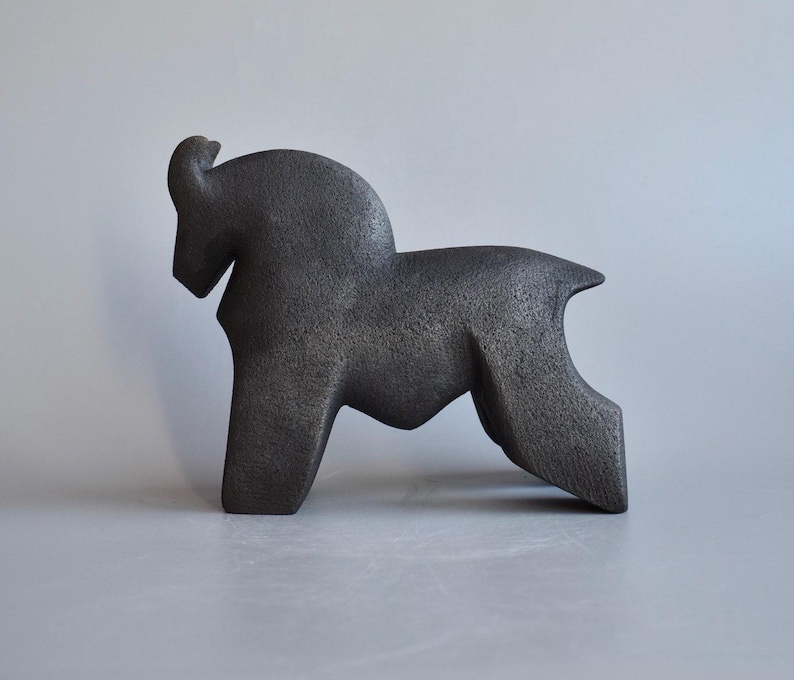 May include: A dark charcoal gray sculpture of a stylized animal with a curved back, a prominent head, and a textured, matte surface. The figure stands on four legs in a solid, abstract form, showcasing a uniform dark finish throughout.