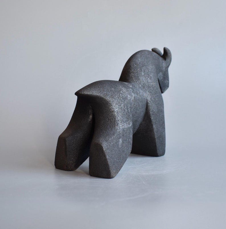 May include: A dark gray, abstract sculpture of an animal, possibly a bull or bison, with a textured surface. The sculpture has a geometric design and is viewed from the side, showcasing its form and shape. The artwork is set against a neutral background.