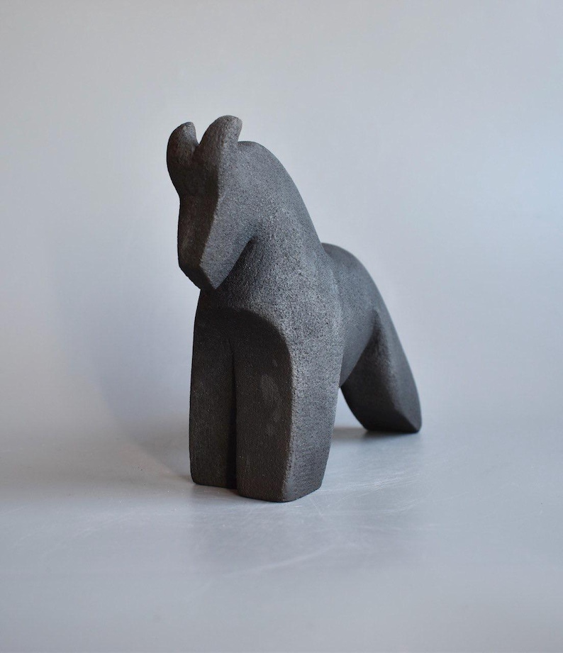 May include: A dark gray, abstract horse sculpture. The sculpture has a geometric design with a curved neck and a forward-leaning posture. The horse's body is solid and blocky, with a smooth, textured surface.