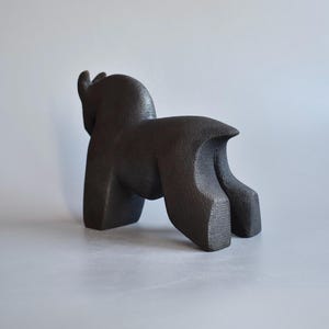 May include: A dark brown, textured sculpture of a four-legged animal with a rounded back, thick legs, and a short tail. The figure is depicted in a standing position, showing a solid, matte surface with a grainy, cast-like appearance.