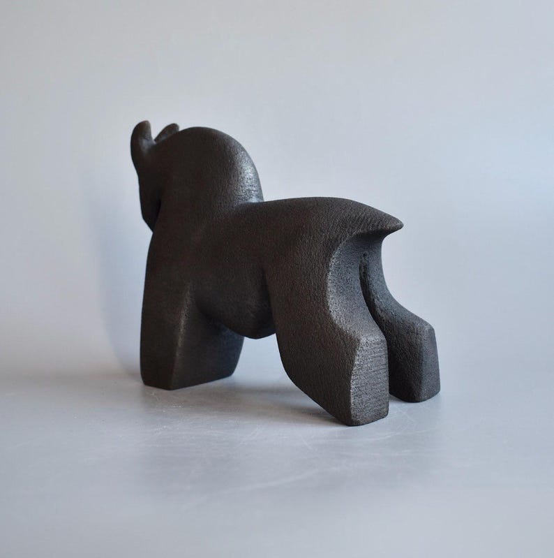 May include: A dark brown, textured sculpture of a four-legged animal with a rounded back, thick legs, and a short tail. The figure is depicted in a standing position, showing a solid, matte surface with a grainy, cast-like appearance.