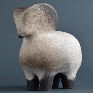 Puede incluir: Escultura de carnero de cerámica con superficie texturizada. El carnero tiene un cuerpo redondeado y patas cortas. La cabeza y las patas son de color gris oscuro, mientras que el cuerpo es de color gris claro. Es un objeto decorativo.