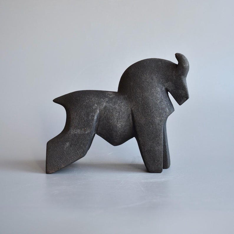 May include: A dark gray, stylized sculpture of a horse in a standing pose. The sculpture has a geometric design with a curved neck and a single horn. The horse is depicted with a simplified form, with angular legs and a streamlined body.