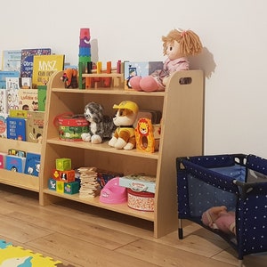 May include: A wooden bookshelf with two shelves filled with books, toys, and stuffed animals. The bookshelf is in a playroom with a blue and white playpen and a colorful foam play mat on the floor.