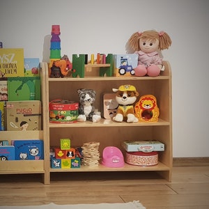 May include: A wooden bookshelf with two shelves, filled with children's books, toys, and games. The top shelf has a doll, a toy truck, a wooden toy, and a puzzle. The bottom shelf has a stack of wooden blocks, a pink potty, a game, and a stuffed animal.
