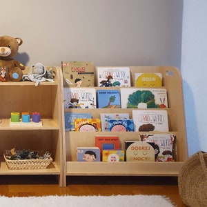 Pu&ograve; includere: Tre librerie in legno con vari giocattoli e libri. La libreria a sinistra ha un giocattolo di legno con una pista per le biglie, un cesto di palline e un contenitore di plastica verde. La libreria al centro ha due orsacchiotti, un giocattolo di impilamento arcobaleno e un giocattolo di impilamento in legno. La libreria a destra ha una collezione di libri per bambini con copertine colorate.
