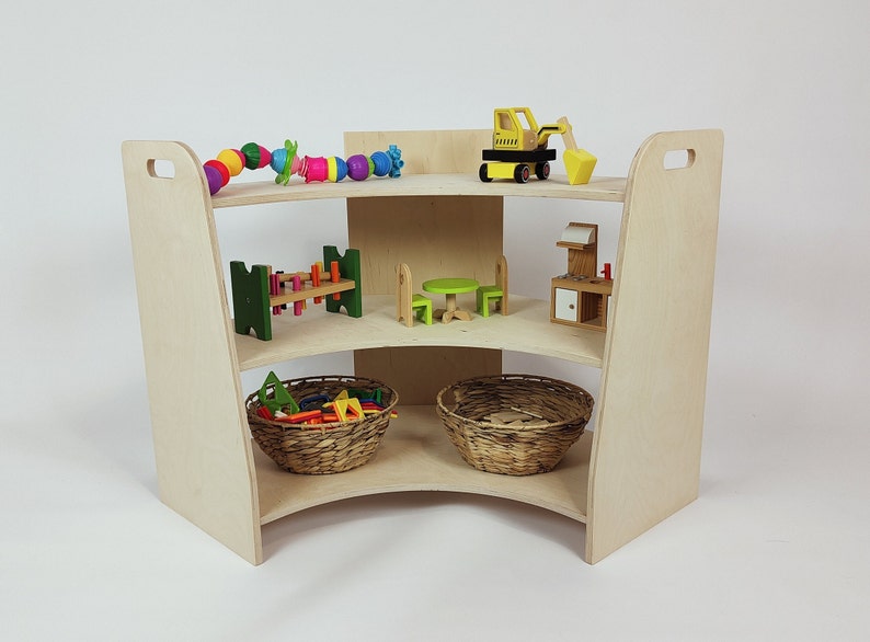 May include: A wooden corner shelf unit with three shelves. The shelves are filled with various toys, including a toy excavator, a toy kitchen, a toy table and chairs, and a toy bridge. There are also two wicker baskets filled with colorful blocks.