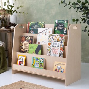 May include: A light-colored wooden bookshelf with multiple shelves, displaying a collection of children's books with colorful covers. The bookshelf has a simple, modern design and is placed against a light green wall.