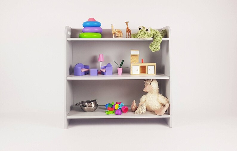 May include: A gray wooden shelf unit with three shelves. The top shelf has a colorful stacking ring toy, two wooden giraffe figurines, and a green frog plush toy. The middle shelf has a purple couch, a pink lamp, a green plant, and a wooden cabinet with drawers. The bottom shelf has a silver colander, a variety of colorful plastic toys, and a white duck plush toy.